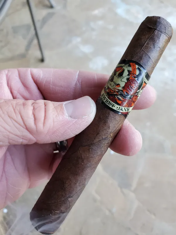 A hand holds a dark brown cigar with a colorful band that says "SWEET JANE" in black letters.